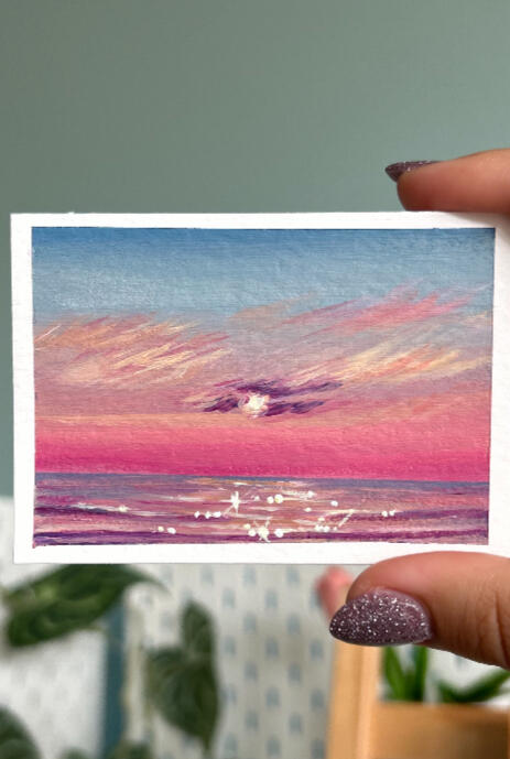 hand holding aceo size painting on paper of a pink sunset over a pink glowing seascape horizon