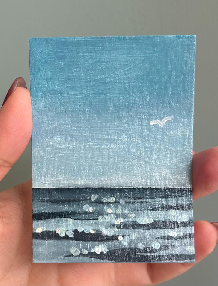 hand holding an aceo sized blue seascape painting on paper. featuring horizon, ripples, blue sky and a seagull silhouette in the sky