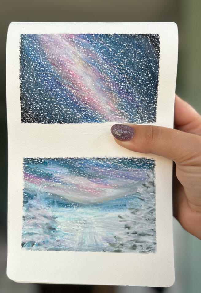 hand holding sketchbook page featuring two mini paintings made with wax pastels. The painting on top, features a dark sky with visible galaxy imagined by the artist, the painting at the bottom represents a snowy landscape with trees and starry night sky